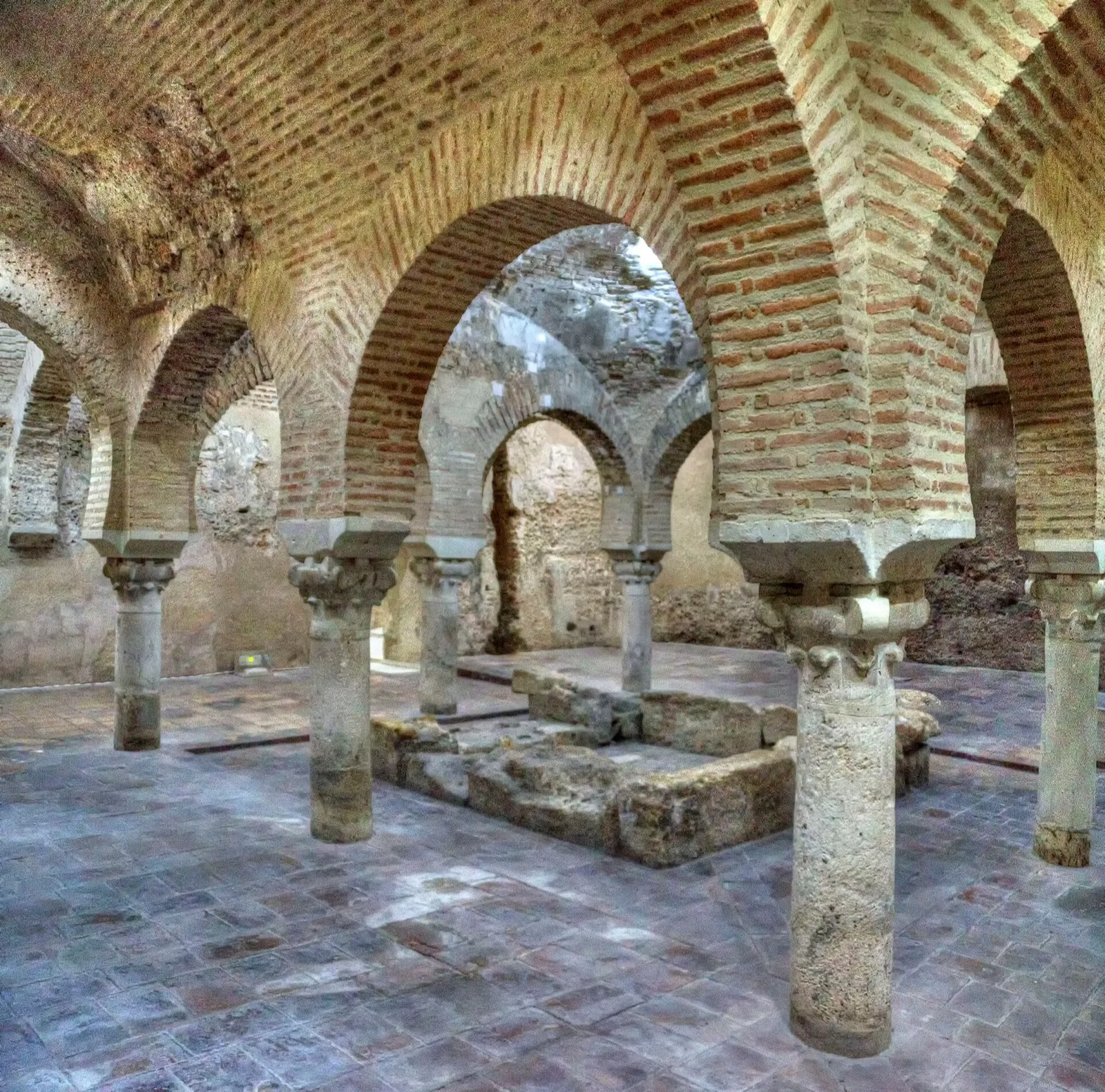 Sala central de los baños árabes de Jaén, con arcos de herradura de ladrillo sobre columnas de piedra y una pila en el centro