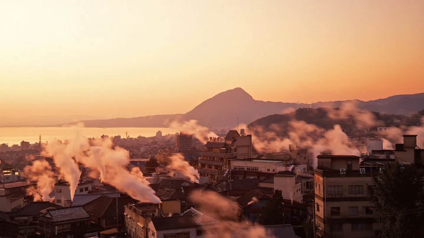 Vapor subiendo de las fuentes termales entre las casas de Beppu, en Ōita, al atardecer, con la bahía al fondo