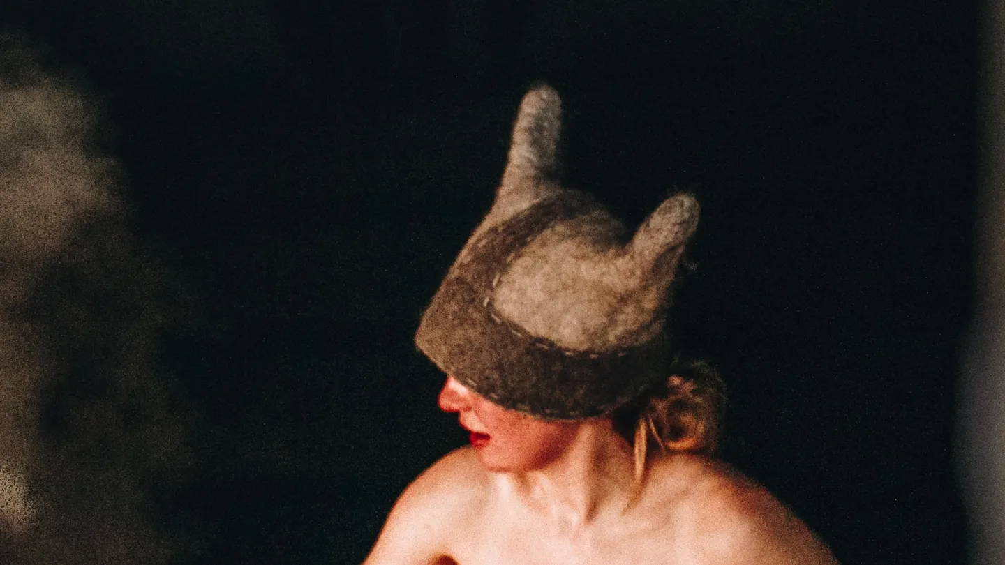 Bañista con un gorro de sauna de fieltro con orejas en un sauna de humo en Estonia, con el vapor subiendo de las piedras al lado