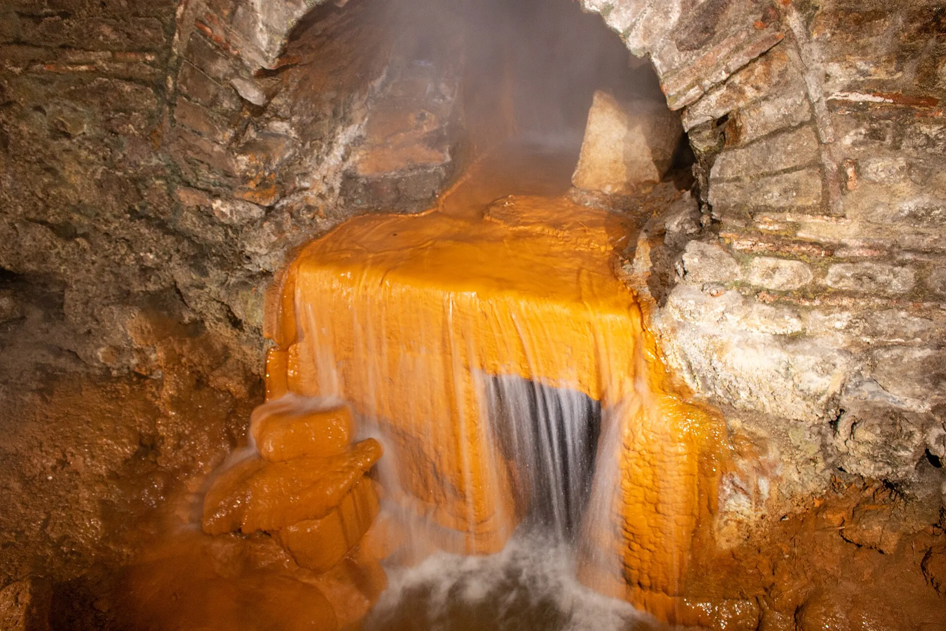 Agua caliente de la Fuente Sagrada de Bath escurriendo sobre piedras teñidas de naranja por el hierro, bajo un arco de piedra de las termas romanas