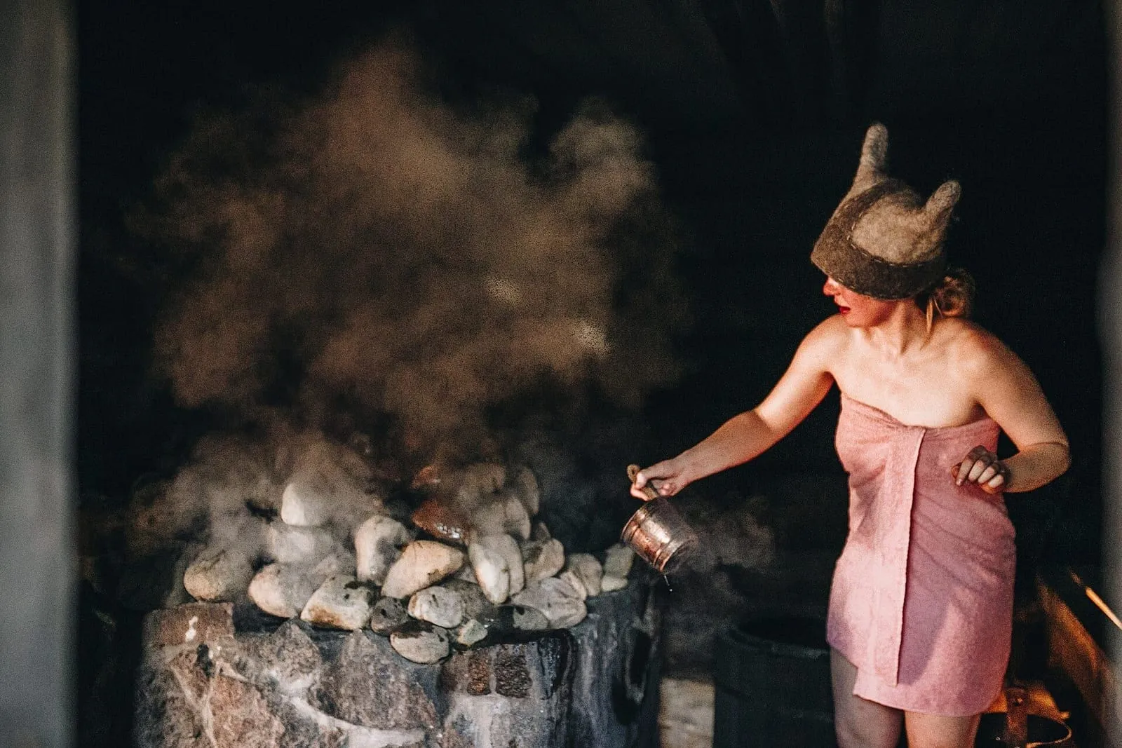 Mujer con toalla y gorro de fieltro echa agua sobre las piedras de un sauna de humo en Estonia, con el vapor subiendo
