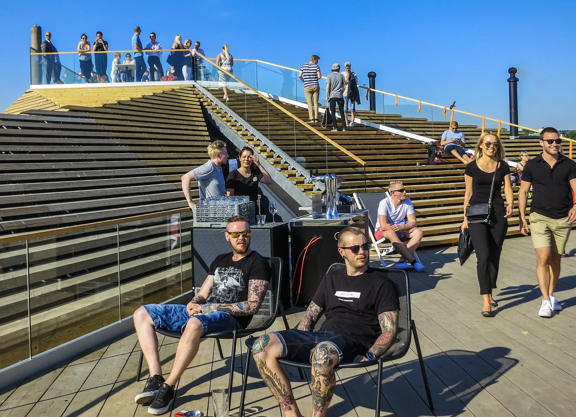 Terraza de madera del Löyly, en Helsinki, con gente sentada en sillas y de pie en la escalera del techo escalonado, bajo un cielo azul