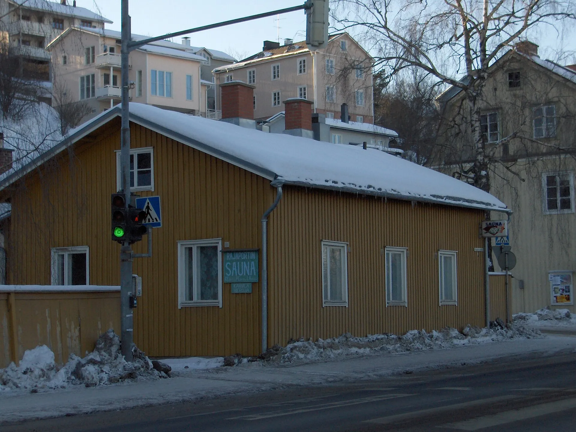 Edificio bajo de madera amarilla en la esquina de una calle nevada de Pispala, en Tampere, con el letrero del sauna Rajaportti en la fachada