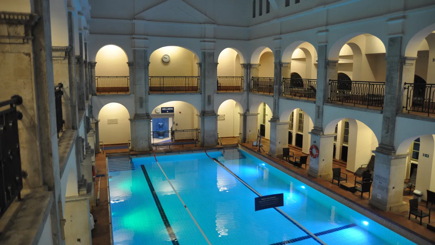 Alberca interior vacía de los Baños Rudas, en Budapest, con agua azul iluminada y galerías de arcos en dos niveles
