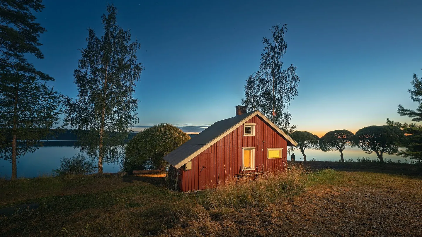 Cabaña de sauna de madera roja a la orilla del lago Ätäskö, en Kitee, Finlandia, con luz encendida al anochecer